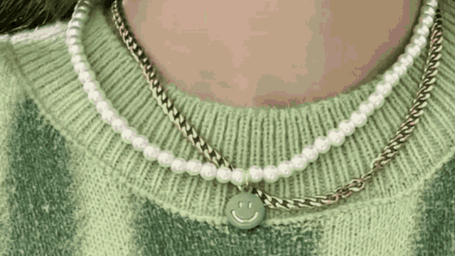 a close up of a person wearing a green sweater and a necklace with pearls and a smiley face pendant .