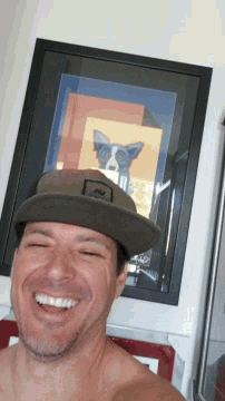a shirtless man wearing a hat with the letter n on it smiles in front of a framed picture of a dog