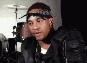 a man wearing a bandana and a black jacket is sitting in front of drums .