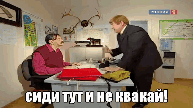 a man sitting at a desk talking to another man with a sign that says russia 1 on it