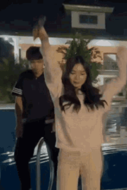 a man and a woman are standing next to a pool at night . the woman is dancing with her arms in the air .