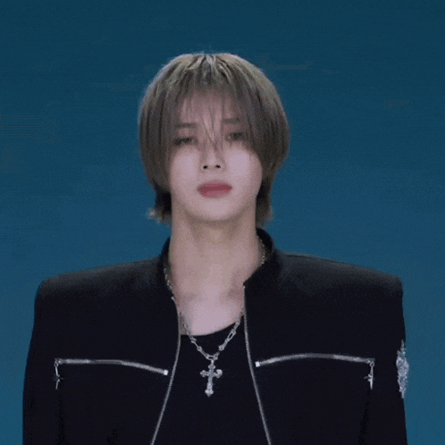 a young man wearing a black jacket and a cross necklace is looking at the camera .