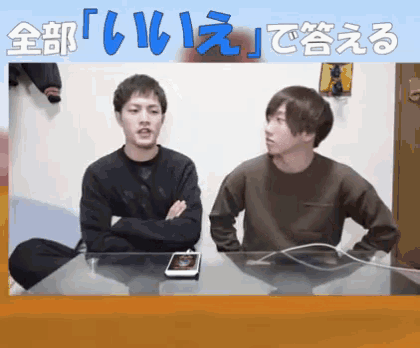 two young men are sitting at a table with their arms crossed and a cell phone on the table ..