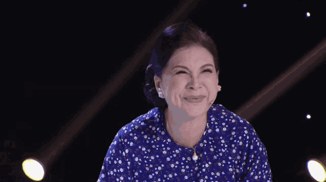 a woman wearing a blue floral shirt and a microphone smiles on stage