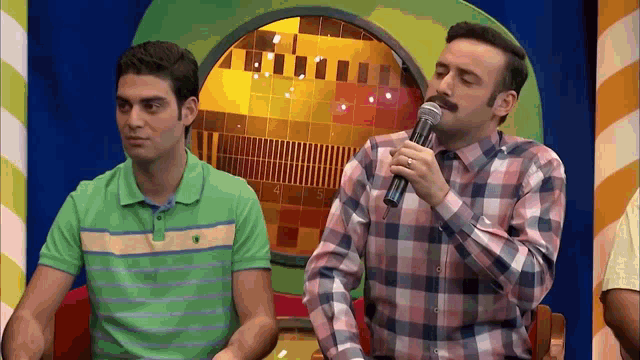 a man with a mustache is holding a microphone while another man looks on