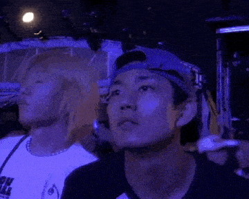 two young men are standing next to each other at a concert . one of the men is wearing a baseball cap .
