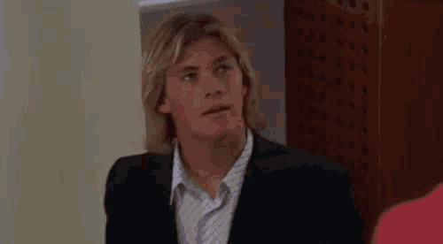 a young man with long blonde hair is wearing a suit and a necklace and looking up .