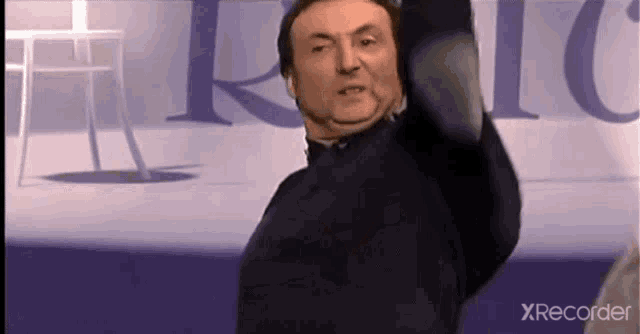a man in a black jacket is standing in front of a purple wall with his arms outstretched .