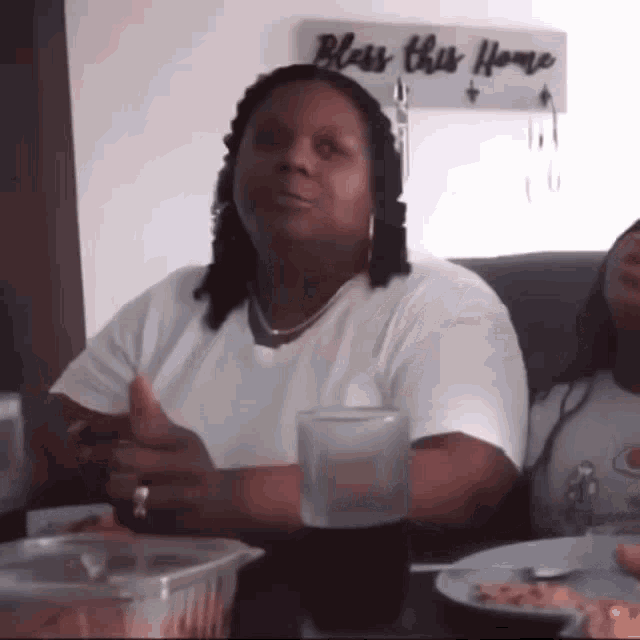 a woman is giving a thumbs up while sitting at a table in front of a sign that says bless this home