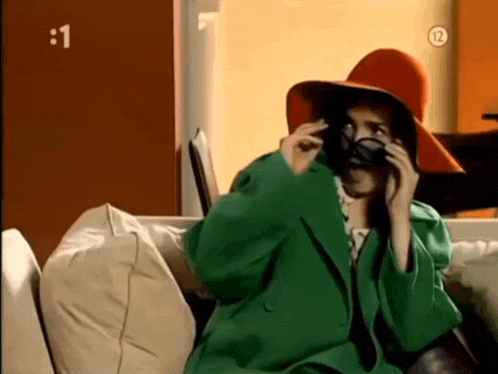 a woman wearing a green coat and a red hat is sitting on a couch talking on a cell phone ..