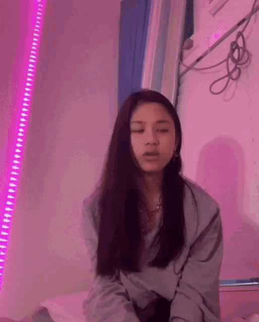 a young woman is sitting on a bed with her eyes closed in front of a pink wall with purple lights .