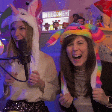 two women wearing unicorn and rainbow hats are smiling in front of a welcome sign