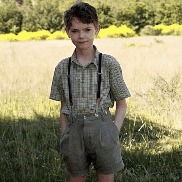 a young boy wearing suspenders stands in a field