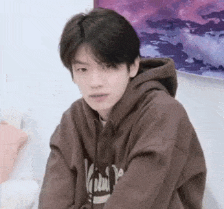 a young man wearing a brown hoodie is sitting in front of a painting and looking at the camera .