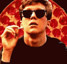a man wearing sunglasses is smoking a cigarette in front of a large pepperoni pizza