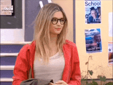 a woman wearing glasses and a red jacket is standing in front of a wall with school posters on it