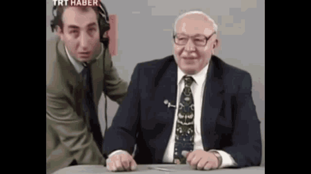 a man in a suit and tie is sitting at a table next to another man with a headset on .