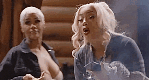 two women are standing next to each other in front of a log cabin smoking a cigarette .
