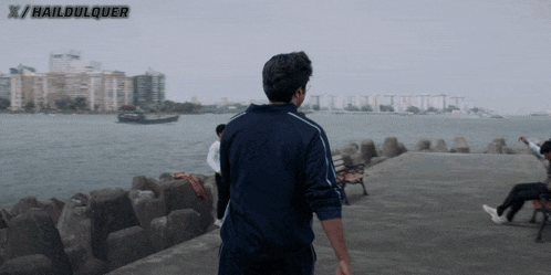 a man in a blue jacket is walking along a pier with haildulquer written on the bottom