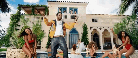 a man in a yellow jacket is standing in front of a mansion surrounded by women in swimsuits .