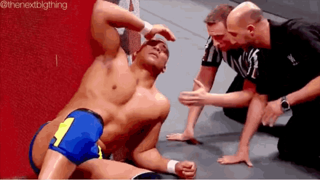 a wrestler is laying on the floor with his head in his hands while two referees look on .