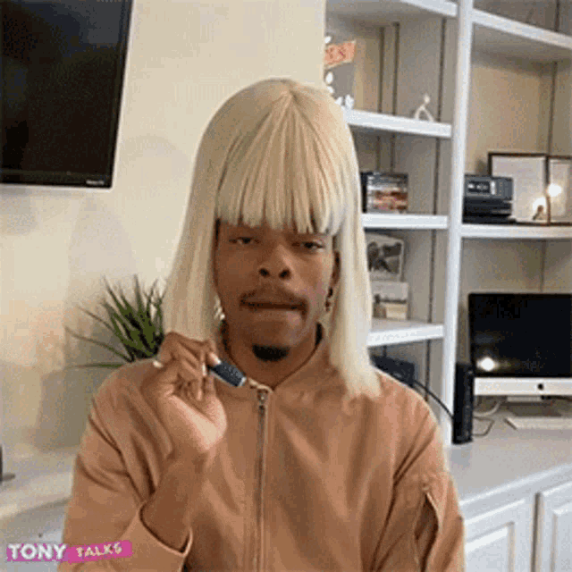 a man wearing a blonde wig and a tan jacket is sitting in a room .