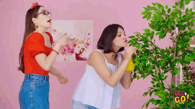 two women standing next to a plant with a sign that says ' a23 go ' on it