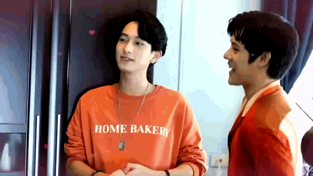 two young men are standing next to each other in a kitchen . one of the men is wearing a home bakery shirt .