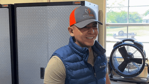 a man wearing a hat that says wolftech stands in front of a fan