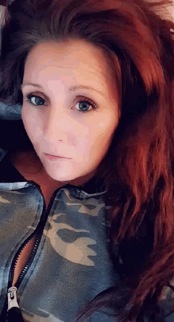 a woman with long red hair is wearing a camouflage jacket