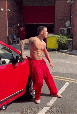 a shirtless man in red pants leans on the side of a red car