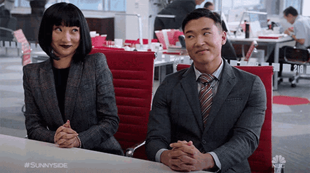 a man and a woman are sitting at a table with their hands folded in front of them in an office .