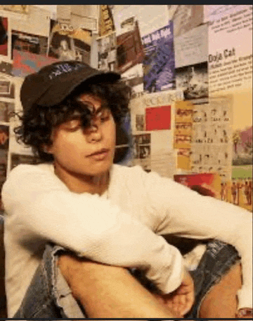 a person wearing a hat and a white shirt is sitting in front of a wall with posters on it .