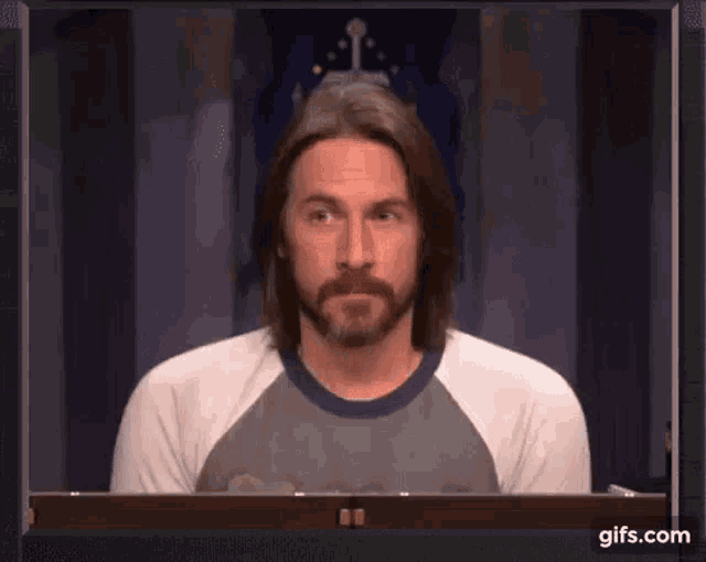 a man with long hair and a beard is sitting in front of a television .