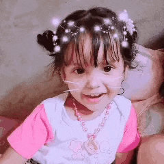 a little girl wearing a pink shirt and a necklace is smiling while sitting on a couch .