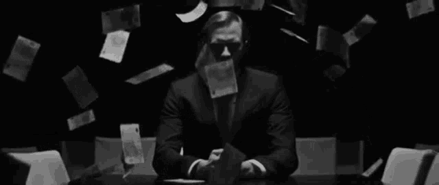 a man in a suit is sitting at a table with money falling from the sky .