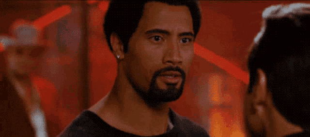 a man with a beard is talking to another man in a dark room