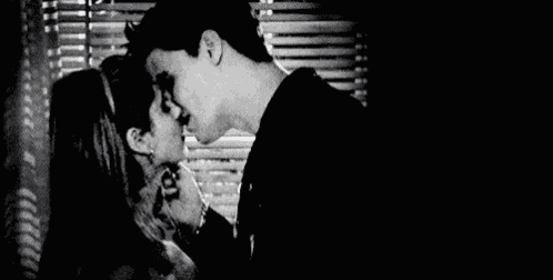 a black and white photo of a man and a woman kissing in front of a window .