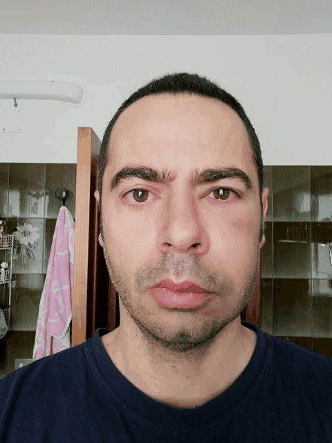 a man in a blue shirt looks at the camera with a pink towel hanging on the wall behind him