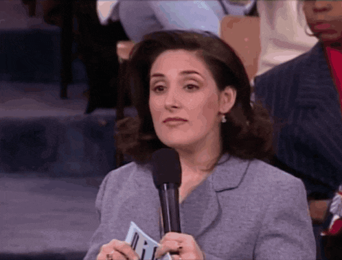 a woman in a suit holds a microphone and a piece of paper that says ' i ' on it