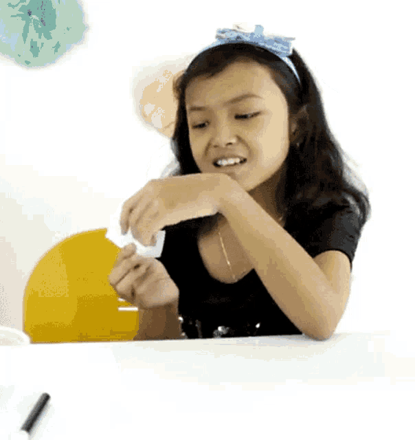 a little girl wearing a blue headband is sitting at a table playing with a piece of paper