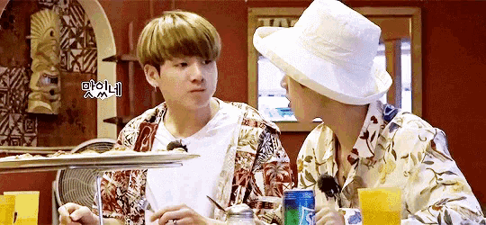 two men are sitting at a table eating food and drinking soda . one of the men is wearing a hat .