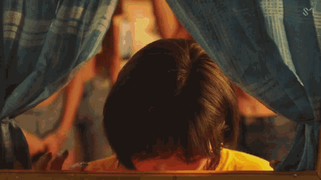 a woman in a yellow shirt is looking out of a window with a blue curtain behind her