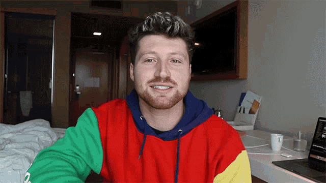 a man in a colorful sweatshirt is smiling in front of a macbook pro