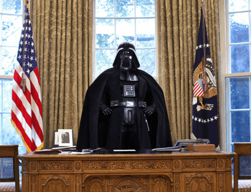 a man dressed as darth vader is sitting at a desk in front of two american flags