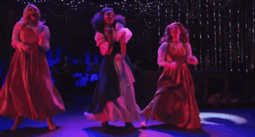 three women in long dresses are dancing on a stage with purple lights behind them
