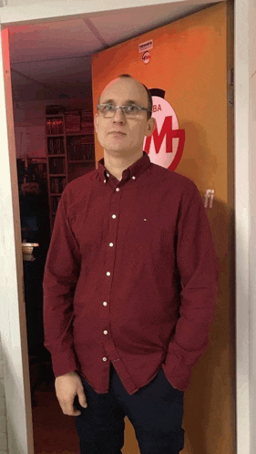 a man in a red shirt and glasses stands in front of a door that has a m on it