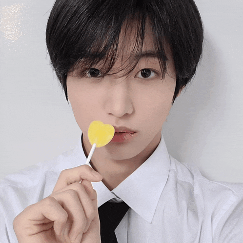 a young man in a white shirt and black tie is holding a yellow heart shaped lollipop in his mouth
