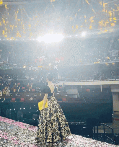 a woman in a floral dress stands on a stage in front of an empty stadium with confetti falling from the ceiling