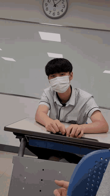 a boy wearing a face mask sits at a desk in front of a clock that shows the time as 7:15